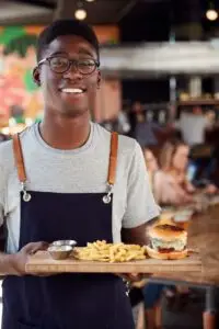 Portrait Of Waiter Serving Food To Customers In Busy Bar Restaurant